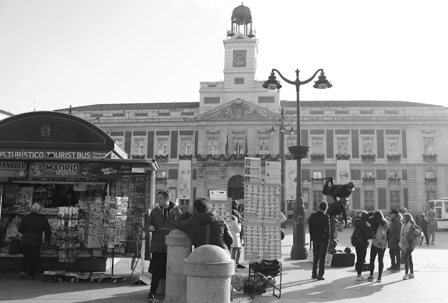 Plac Puerta del Sol w okresie przedświątecznym, pełen ludzi i ulicznych sprzedawców loterii bożonarodzeniowej. W tle budynek siedziby regionalnych władz, ze znajdującą się na jego osi cetralnej wieżą z zegarem. Zdjęcie czarno-białe. 