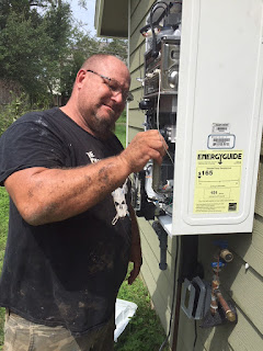 A plumber stands next to a 3 foot by 4 foot white metal box mounted on the outside wall of a house. He uses a small tool to fix the guts of the machine.