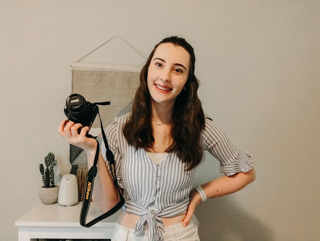 Madalyn smiling and holding a Nikon D3500 camera while standing in front of a woven wall hanging.