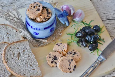 Ein Foto mit Olivenbutter in einem Porzellan-Fass. Daneben zwei Scheiben helles Brot. Rechts liegen auf einer hellen Platte mehrere schwarze Oliven und Kräuter.