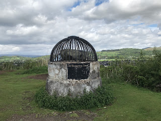 A photo of the Beheading Stone in Stirling. It is a large grey stone that is sitting on a round stone plinth and it is enclosed in an iron cage.  There is a plaque on the front that reads - Protected by the public at the instance of the Stirling Natural History and Archaeological Society 1887.  Photograph by Kevin Nosferatu for the Skulferatu Project.