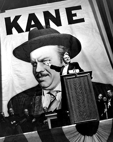 a small Orson Welles at a podium, raising his hand in front of a huge poster of Orson Wells labeled "Kane"