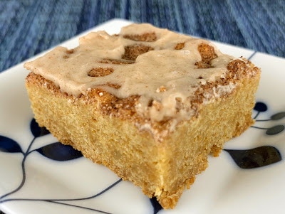 A cinnamon sugar blondie, topped with cinnamon icing. Photographed on a white plate with leaves.