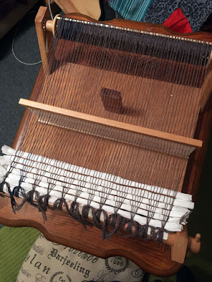 A shot from above of a rigid heddle table loom made of light natural wood, with mostly grey and some teal warp threads wound onto the back beam and tied to the front beam. The heddle is propped on edge in the center of the loom, and six rows of crushed toilet paper are woven into the base of the warp.