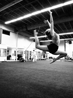 black and white photo of a dark haired fair skinned person doing a leap. their back foot is up by their head and their front knee is bent at an acute angle