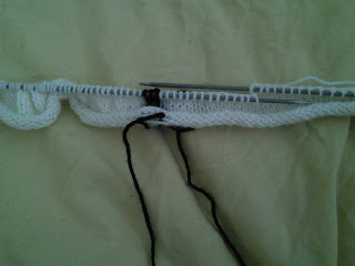 a square-in-progress, knit in stocking stitch with white yarn, with a small bit of black colourwork in the centre.