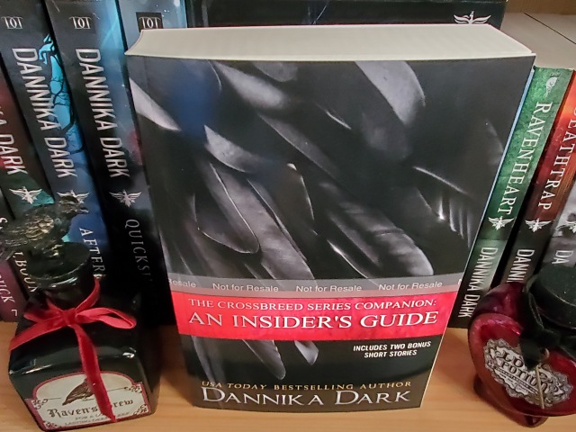 Insider's Guide paperback sitting on bookshelf next to other Crossbreed books. Decorative bottle beside it with a Raven on top of the cork. Heart-shaped Love Potion bottle on opposite side.