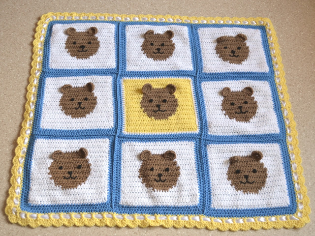 A square baby blanket comprised of 9 squares, all white except for the centre square which is yellow. Each square has a blue border and a tan teddy bear face in the middle. The teddies have three dimensional ears stitched onto the surface of the blanket. The outside border of the blanket is crocheted in yellow with a row of filet crochet and ending with a scalloped edge. White satin ribbon is woven through the filet spaces.