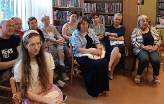 Zasłuchani uczestnicy spotkania autorskiego z Natalią Szyszkowską, na tle okna i regału z książkami.
