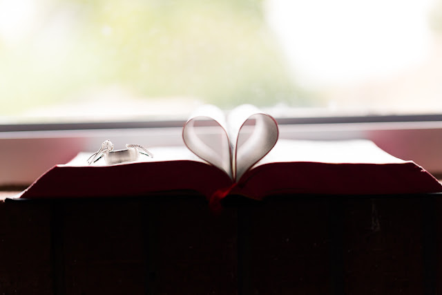 Her solitaire engagement ring and diamond wedding band rest on his beveled edge brushed finish white gold wedding band.  The rings sit atop a bible, whose inner pages have been folded inward to create a heart shape.  The scene is backlit from a large window in the Southern Oklahoma Chinese Baptist Church, located in Norman, OK.