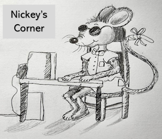 Nickey, a blind mouse, is seated at a table in Nickey's Corner with his front paws on a computer keyboard. He is wearing a short sleeve shirt and shorts with a bowtie and sunglasses. The tip of his tail is bandaged.