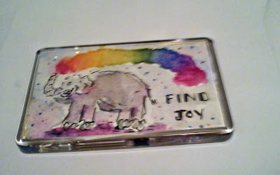 Watercolour fridge magnet in which a happy grey elephant is spouting rainbow dust from its trunk, and the message Find Joy, is hand-lettered i black.