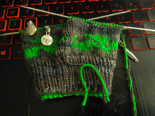 A double-knit glove live on double-pointed needles  Yarn is brown, with a dragon motif around the cuff knit in green.  Two stitch markers with charms that read "Faith" are clipped into the work, and a stitch marker with a silver feather charm is at the side of the work.