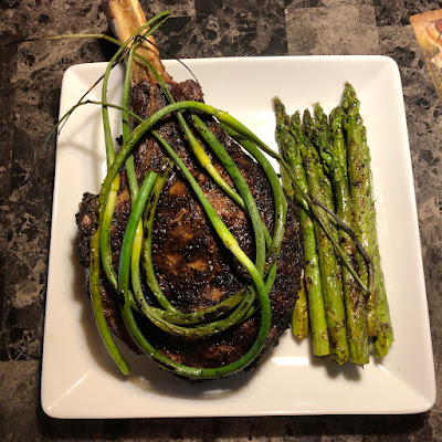 Prime tomahawk ribeye grilled Pittsburgh rare, topped with grilled garlic scapes along with grilled asparagus