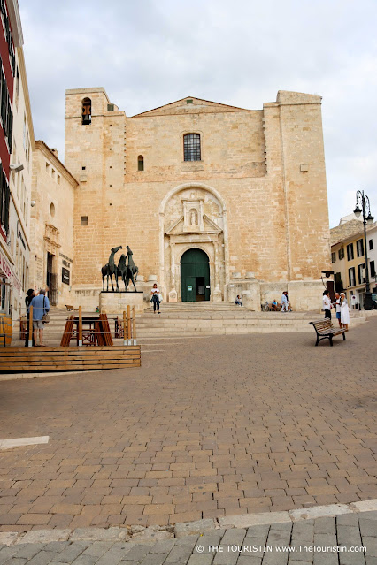 The statue of three horses next to the front entrance of a large sandstone cathedral on a square frequented by only about ten people.