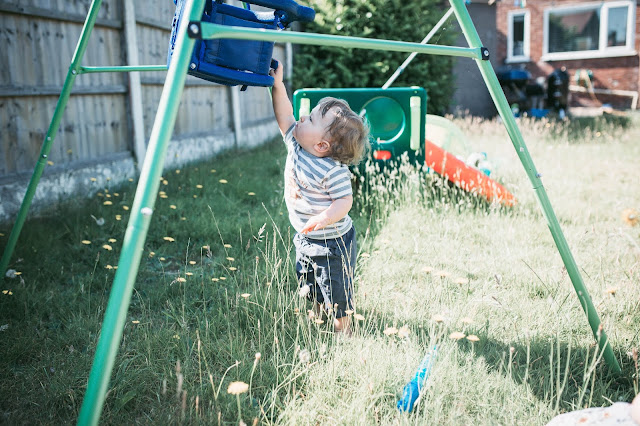 blonde toddler boy standing in a garden and reaching up to touch a blue swing