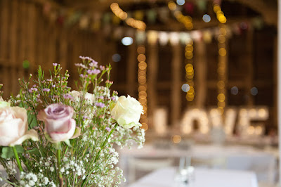 A floral centre piece of purple and yellow roses on a white tablecloth in a rustic old barn.