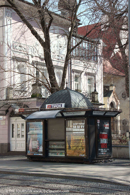 An art-deco-style kiosk on a cobblestone road with tram tracks in front of a grand white period-style mansion.