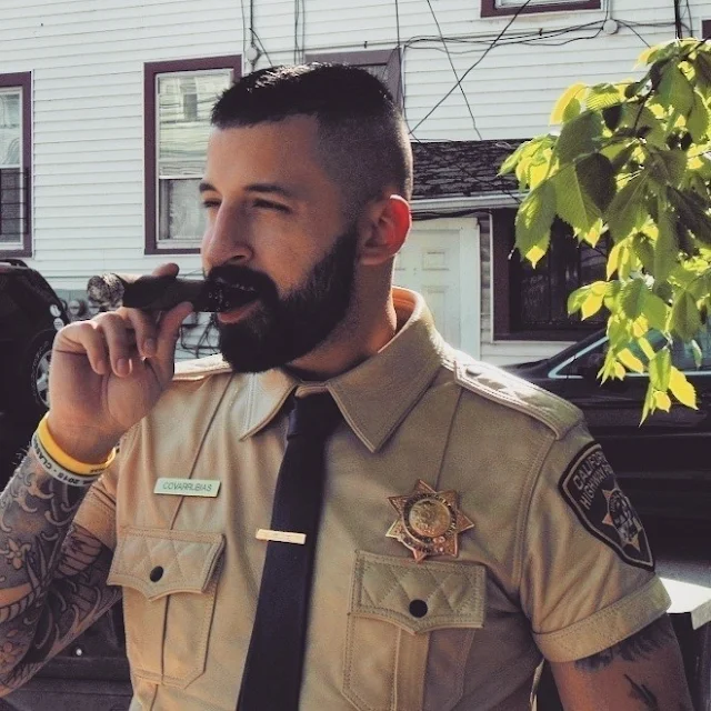 Leatherman wearing a tan cop uniform shirt with tattoos on his arms and the cigar in his mouth in somebody's backyard