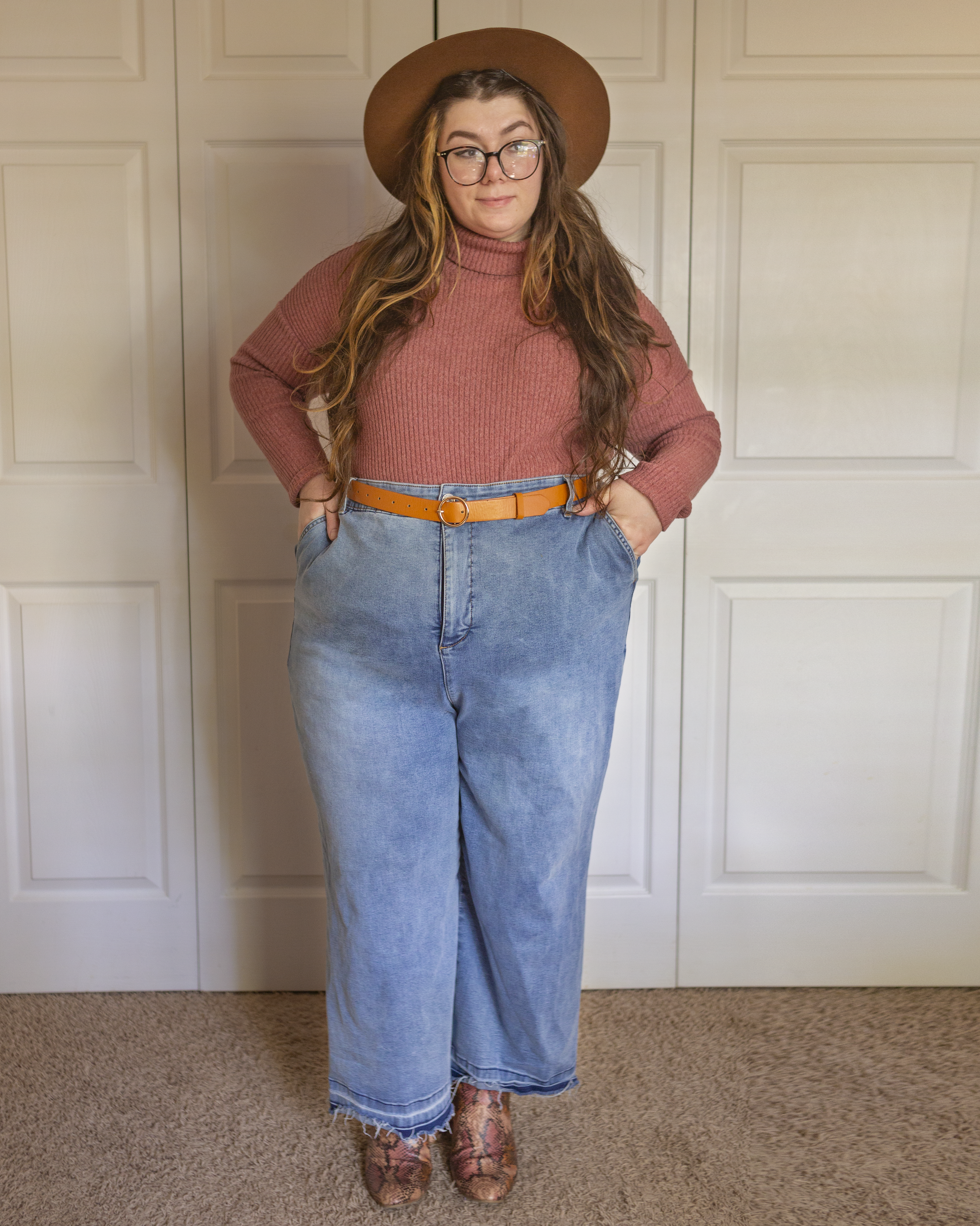 An outfit consisting of a cognac brown panama hat, a muted pink lightweight turtleneck sweater tucked into medium wash wide leg jeans and muted pink, brown and tan snake skin boots.