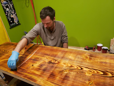 Image shows Max pouring a layer of clear apoxy over a large slab of wood which has been lightly burned using a tiger torch to highlight the grains of the wood. Max is wearing surgical gloves and distributing the apoxy in one even layer across the slab of wood.