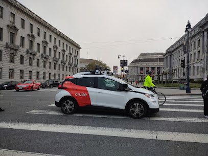 Photo of a red and white self-driving taxi cab stuck in a pedestrian crosswalk, with Davies Symphony Hall and the War Memorial Opera House in the backgroud, and SF City Hall on the right.