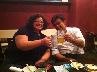 Woman with curly hair and glasses wearing a short sleeve black polo style shirt holding an empty beer stein touching in a cheers motion with a man in a white shirt and black tie holding a nearly full beer stein