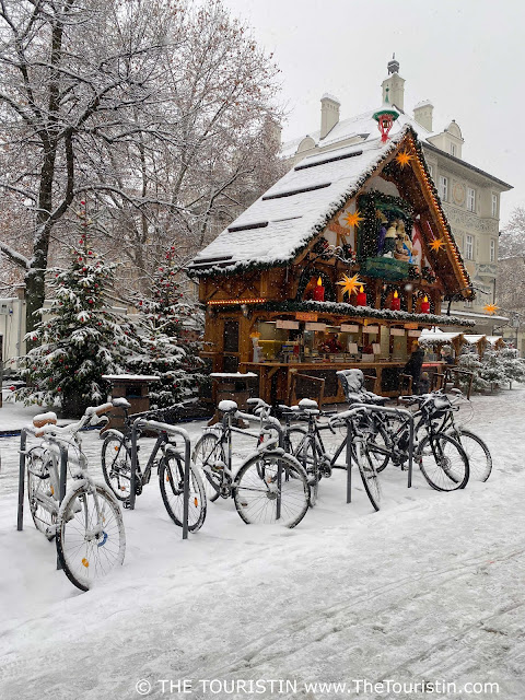 A row of six bicycles parked in front of a wooden stall selling Christmas delicacies and fir trees decorated with red baubles, all covered in a proper dusting of snow.