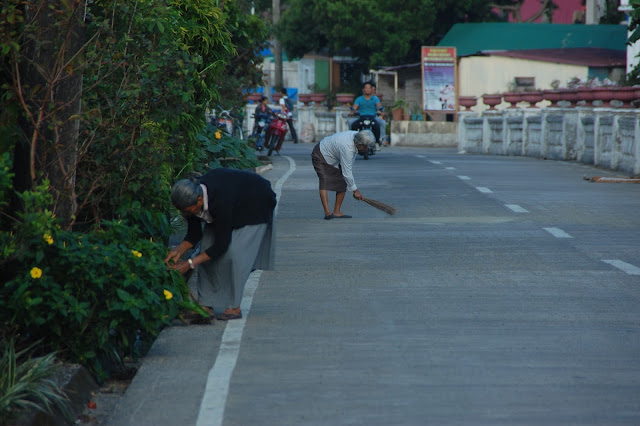 Old ladies sweeping and pruning along the national road of Batan Town, Batanes while younger men go off to work