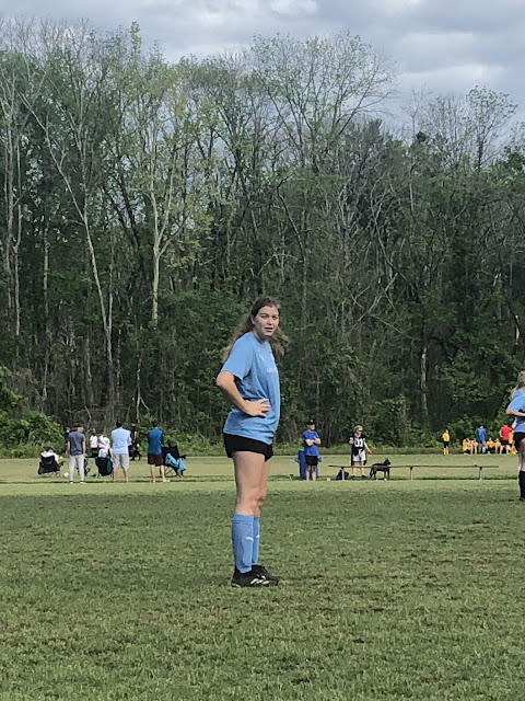 Elizabeth on the soccer field getting ready to start the game. Her hands are on her hips and she is staring in our direction as we sit on the sidelines. She is wearing a baby blue jersey.