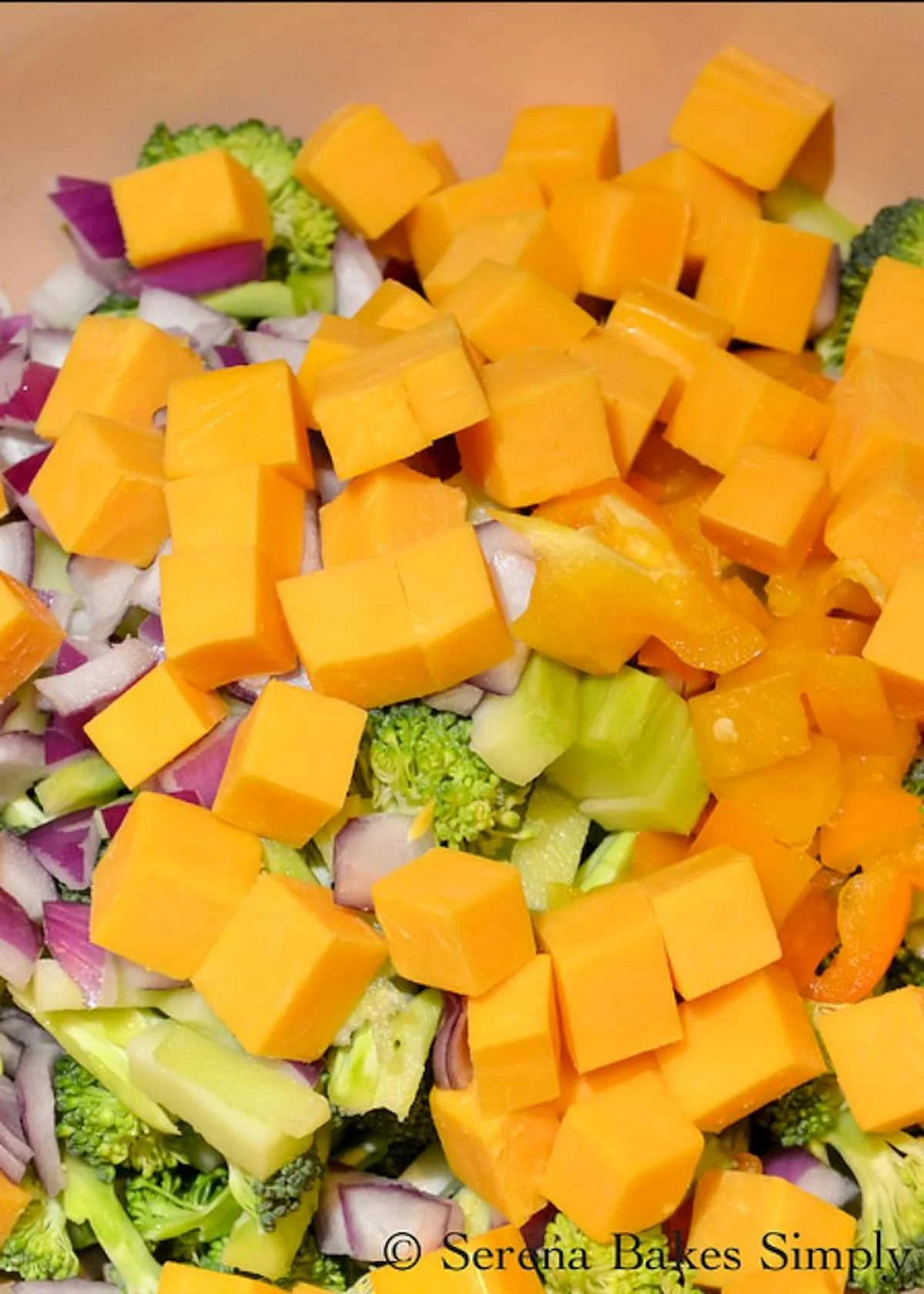 Cubed Cheddar Cheese added to Broccoli, Diced Red Onion, and Diced Red Pepper in a bowl.