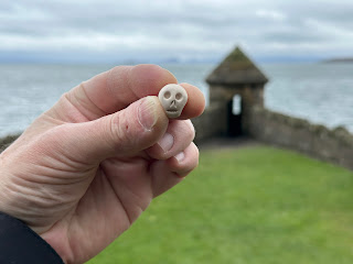 A photo of a hand holding up a small ceramic skull (Skulferatu #126) with a small stone tower in the distance.   Photo by Kevin Nosferatu for the Skulferatu Project.