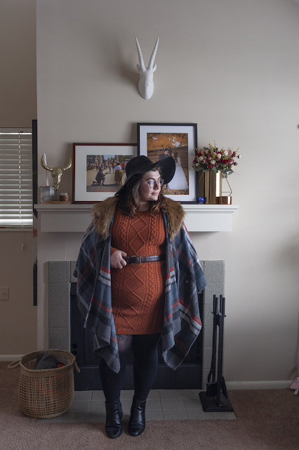 An outfit consisting of a wide brim floppy hat, plaid poncho, orange sweater dress and black ankle boots.