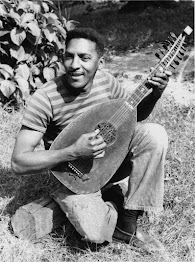 A photo of a smiling Rustin (in his late 20s), outdoors, sitting on a log, strumming a mandolin