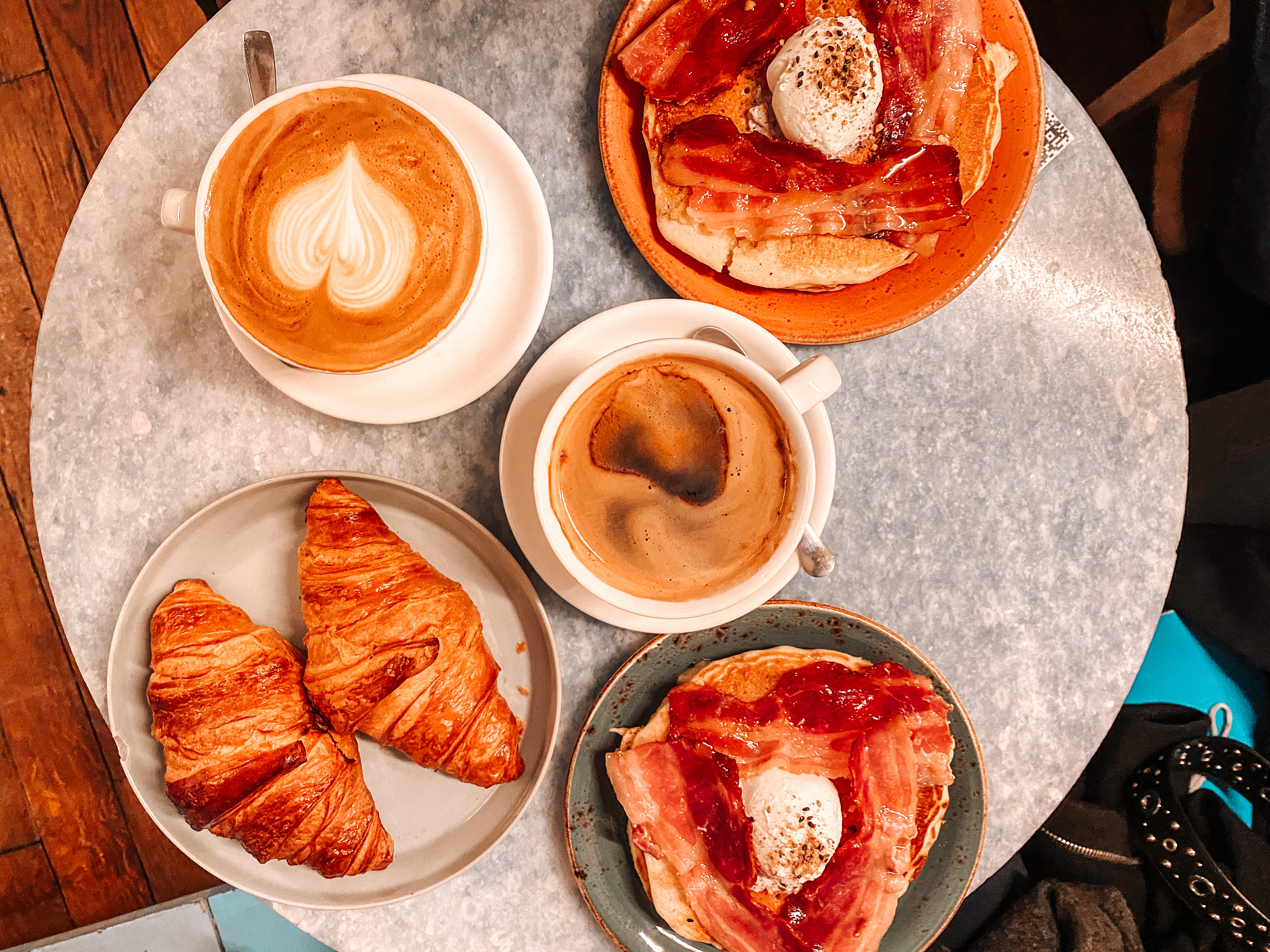 en bas à gauche deux croissants dans une assiette bleue au dessus deux cafés et à droite deux assiettes avec des pancakes et du bacon disposé dessus