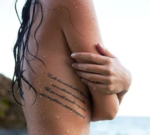 chica en la playa, esta mojada y lleva un tatuaje de frases en las costillas