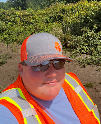 •	Photo shows Beth Blankenship who is wearing sunglasses and an orange with grey baseball cap that features the “WSDOT” logo in orange on the front. Beth is also wearing a bright orange, gray and yellow safety vest with a gray shirt.
