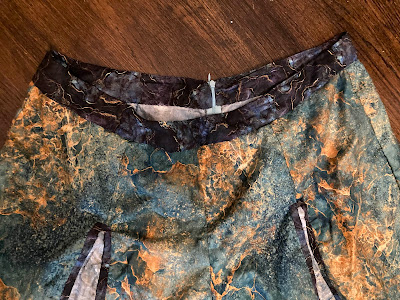 A close-up of the finished waistband for a pair of palazzo pants. The waistband is purple, the main fabric is green and yellow/orange, and the partially visible panels in the legs are grey with purple borders