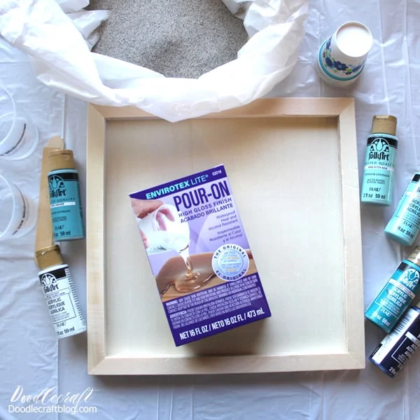 Supplies needed for ocean resin paint pour: Wood canvas, envirotex lite pour on high gloss resin, acrylic craft paint, mixing cups, stirring sticks, and beach sand.