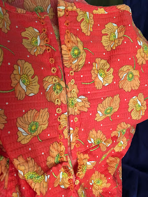 A close photo of the back edges of a coral sundress with large, scattered orange poppy motifs. Eyelets worked in thick variegated orange-and-yellow thread run down either edge; the color shifts are slightly offset from each other. 