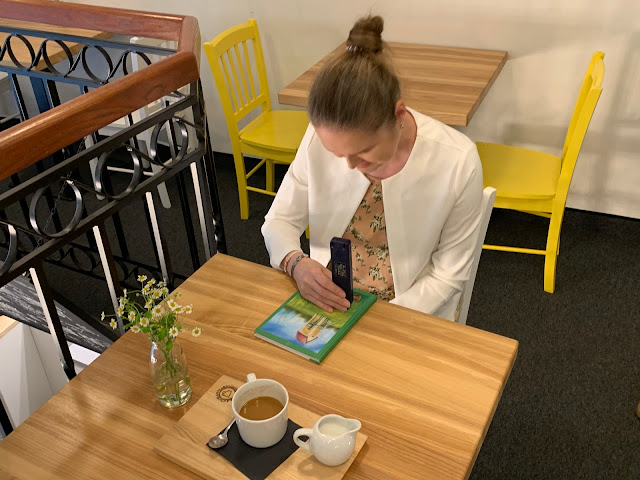 Linda sedí u stolku v kavárně, před sebou má knihu a pomocí colortestu zkoumá barvy malby na titulní straně. Na stolku stojí květina ve váze a hrnek s kávou a konvička s mlékem.