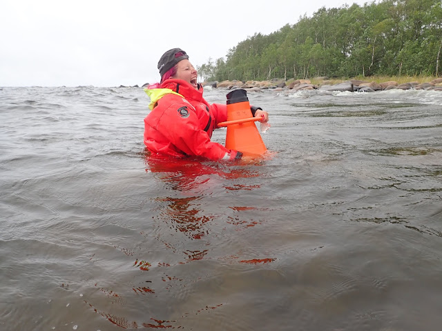 Suvi nauraa vyötäröään myöten meressä seisoessaan pelastautumispuku päällä ja vesikiikarit kädessä. 