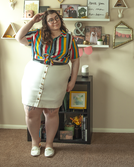 An outfit consisting of a multi colored rainbow inspired button down tie up crop top tied above a white button down midi skirt and white mules.