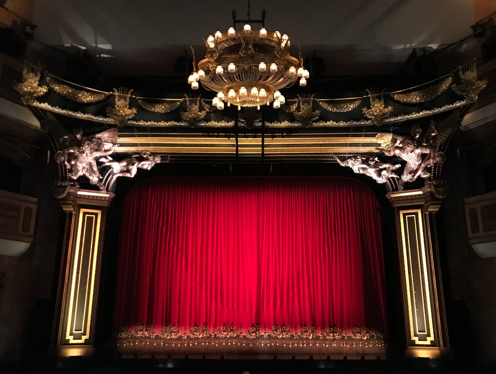 A photo of a theatre, with it's red curtains drawn, two squared coloumns either side with carved figures at the top, and a chandelier hanging from the ceiling at the top of the photo.