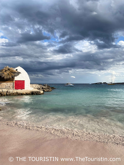 A tiny white upside-down boat-shaped fisherman's cottage with a bright red wooden door on a small sandstone wharf on a golden sandy beach with turquoise water under a darkening sky.