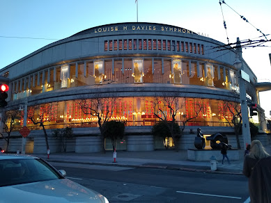 Photo of Davies Symphony Hall, SF; a multistory white building with huge plate glass windows, seen at dusk at a corner angle