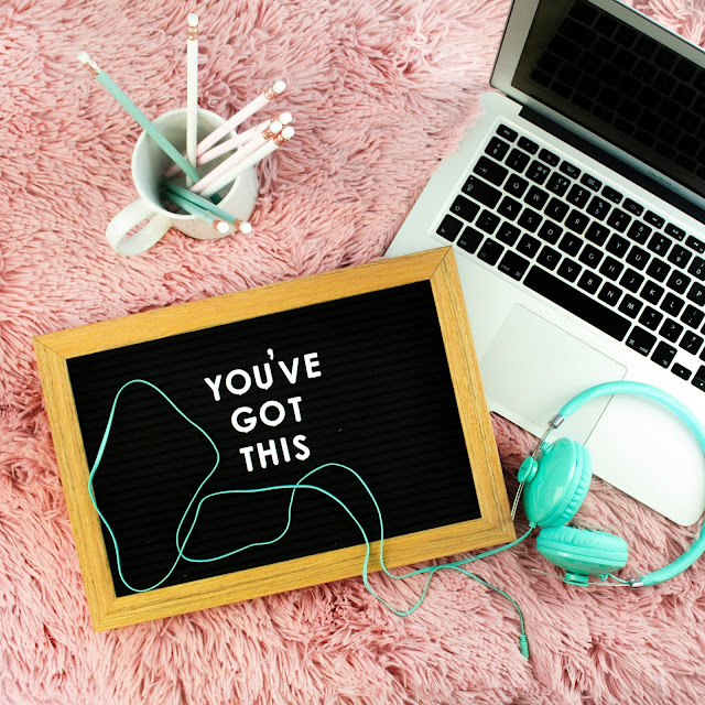 Laptop on a pink rug, next to a pot of pens, headphones and a board that reads 'you got this'