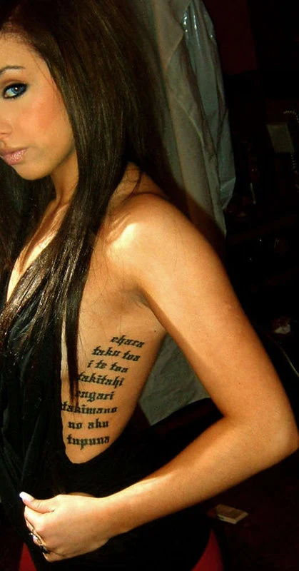 Chica morena de pelo largo, con vestido negro , vemos un tatuaje con frases en las costillas