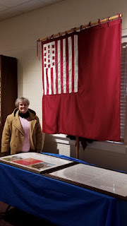 Woman in tan coat stands to left behind a table, there is a red and white Rev War era flag hanging from the ceiling