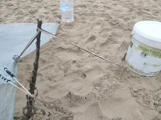 Edge of a tarp, fixed with a stick, string and bucket in the sandy ground of a beach